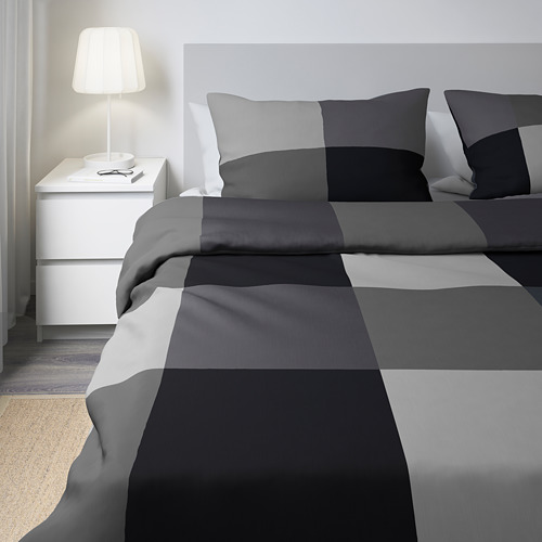 Conjunto de ropa de cama gris BRUNKRISSLA con lámpara, cama grande contra pared blanca. Edredón y fundas de almohada en cuadros grises claros, medios y oscuros.