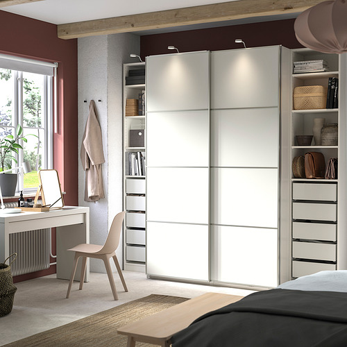 Dormitorio moderno con armario PAX blanco, puertas correderas y estantes organizados. Decoración neutra con escritorio y silla, luz natural por la ventana.