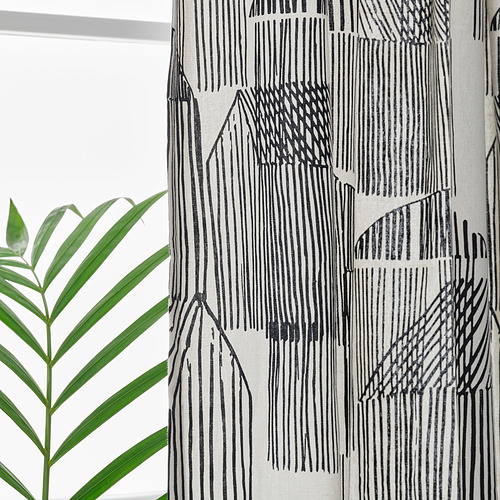 Las cortinas estampadas en blanco y negro cuelgan de una ventana, proporcionando privacidad y filtrando la luz. A la izquierda se ve una hoja verde de la planta.