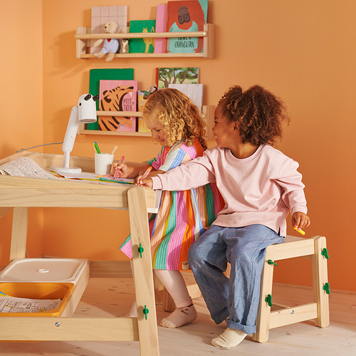 Niños en escritorio ajustable; uno dibuja, otro observa. Paredes naranjas, libros.