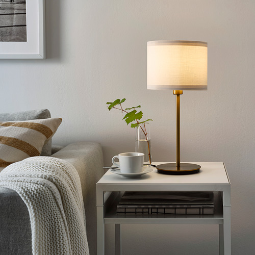 Acogedora sala de estar con lámpara SKAFTET sobre mesa pequeña, sofá gris y planta.
