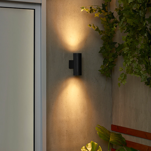 La lámpara de pared GRÖNSPRÖT negra ilumina la pared. Diseño elegante y compacto; emitiendo luz cálida. Rodeado de plantas; más pequeña que la puerta adyacente.