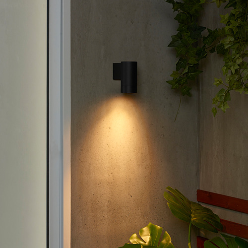 Pequeña luz exterior GRÖNSPRÖT negra, diseño moderno, montada en lo alto de la pared, proyectando un cálido resplandor hacia abajo, rodeada de exuberantes plantas verdes.