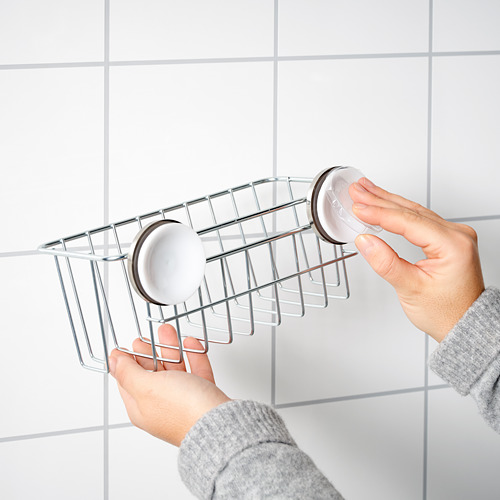 La persona instala la cesta de ducha KROKFJORDEN en la pared con ventosas, la cesta de metal gris tiene capacidad para dos jaboneras blancas redondas.