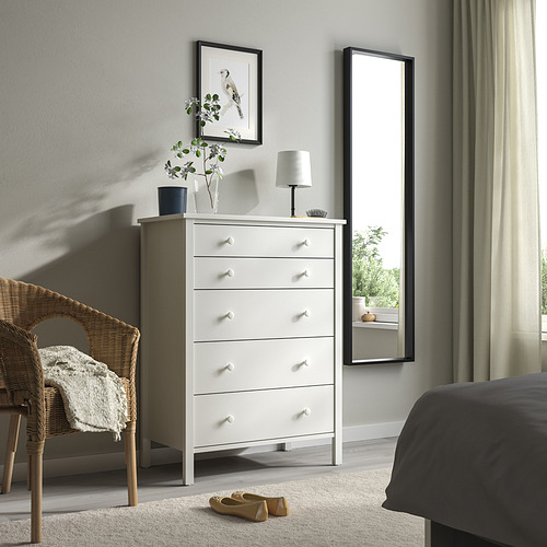 Dormitorio minimalista con baúl alto KILBERGET blanco, lámpara, jarrón, silla de mimbre y espejo grande.