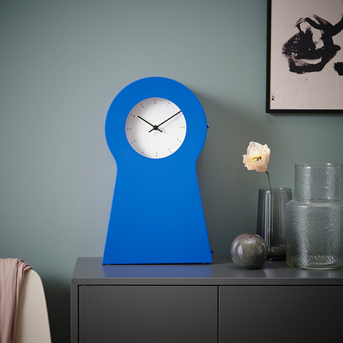 El reloj azul brillante IKEA PS 1995 se alza sobre un estante oscuro, su forma curva única contrasta con un marco de pared simple y una delicada flor.