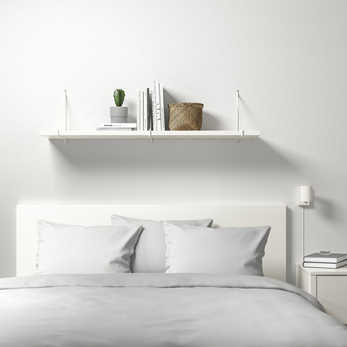 Dormitorio minimalista con estantería de pared BERGSHULT que sostiene libros, plantas, cestas. Líneas blancas limpias.