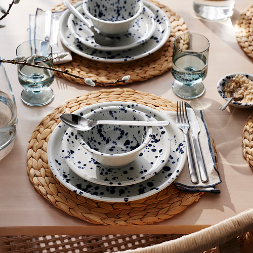 Mesa puesta con platos blancos moteados de azul, colocados sobre manteles individuales tejidos. Los utensilios y la cristalería acompañan, creando una configuración de comedor elegante e informal.