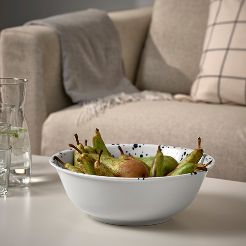 Un cuenco de frutas verdes se encuentra sobre una mesa junto a una jarra de vidrio. Detrás, un sofá beige con almohadas.