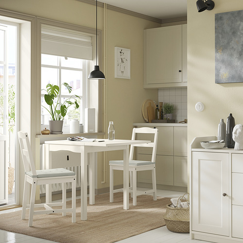 Cocina luminosa con juego de comedor blanco, pared verde, gran ventanal, planta, lámpara colgante y alfombra beige.