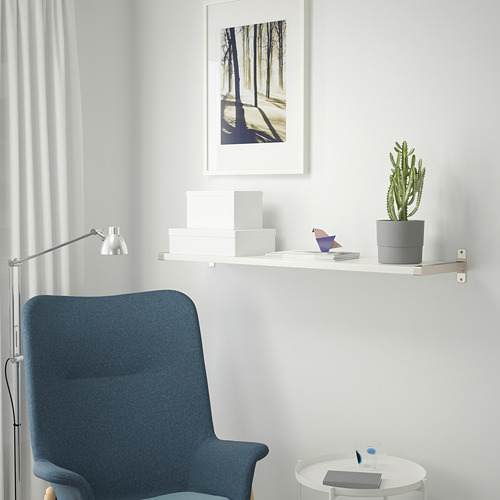 Un espacio de trabajo moderno con una silla BERGSHULT azul, una estantería blanca que sostiene un cactus, libros y un cuadro enmarcado. El estante se monta mediante un soporte metálico.