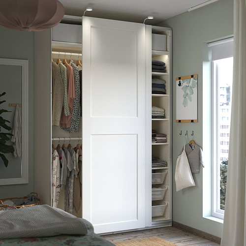 Un armario blanco IKEA PAX con puertas correderas, que contiene estantes, perchas y cestas de malla, organizadas con ropa.