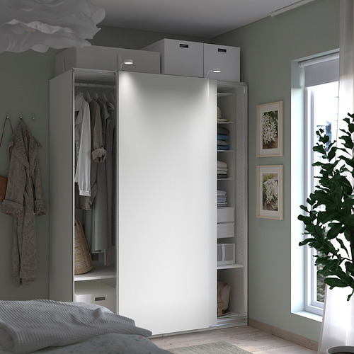 Dormitorio moderno con armario blanco abierto que muestra ropa y estantes, junto a una cama, arte mural enmarcado y una gran planta en maceta junto a una ventana.