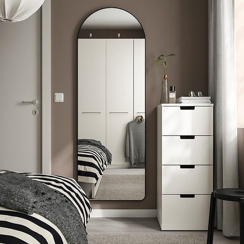 Dormitorio moderno con espejo arqueado LINDBYN, gran armario blanco, cama con marco negro, cajones blancos y taburete. El espejo y el armario dominan el espacio.
