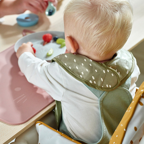 Bebé en silla alta con babero floral, comiendo fruta de un plato gris.