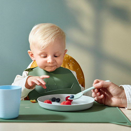 Un niño pequeño con un babero de silicona verde con un bolsillo amarillo, comiendo bayas de un plato blanco con un adulto alimentándolas con una cuchara azul claro.