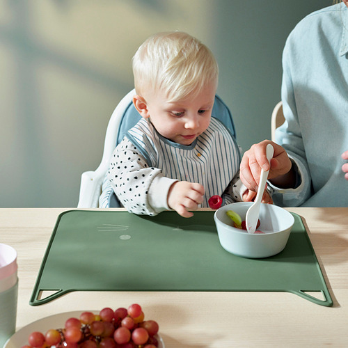 Niño en silla alta comiendo de un cuenco con cuchara. Mantel individual de silicona verde protege la mesa, tiene diseño de gato. Asistencia de adultos.