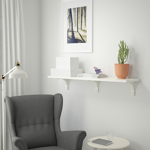 Una habitación minimalista con un sillón gris, una estantería blanca BERGSHULT montada en la pared con libros, una pequeña planta de cactus y un cuadro del bosque enmarcado.