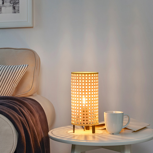 Acogedora lámpara de mesa SAXHYTTAN con pantalla de papel tejido junto al sillón, que emite una luz cálida sobre una pequeña mesa con taza de café.