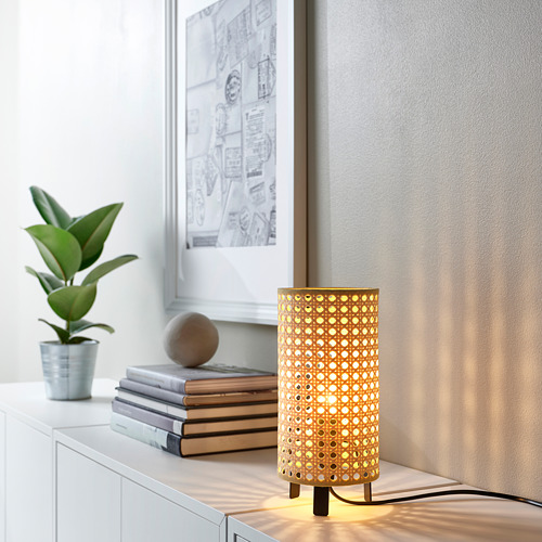 La lámpara SAXHYTTAN con pantalla de papel tejido brilla cálidamente, situada en un estante junto a libros y una planta. Pequeño, con diseños intrincados, que emite una luz suave.