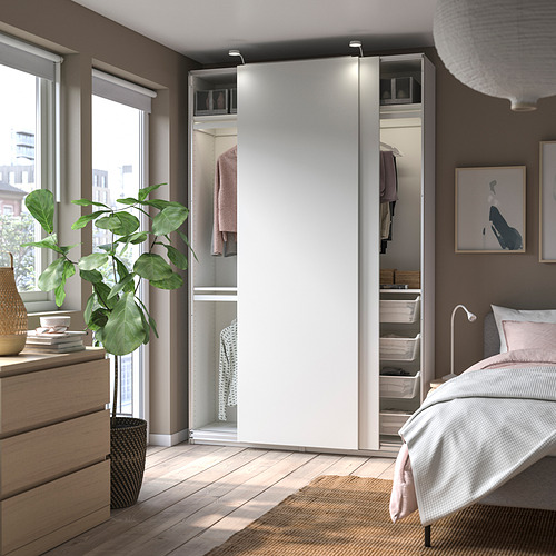 Un moderno armario blanco con puertas correderas en un dormitorio. Lámpara colgante, planta grande, cama y amplia ventana con vista.