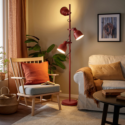 La lámpara de pie HEKTAR en rojo se alza en un acogedor salón, iluminando un sillón gris y un sofá beige, complementando la decoración rústica.