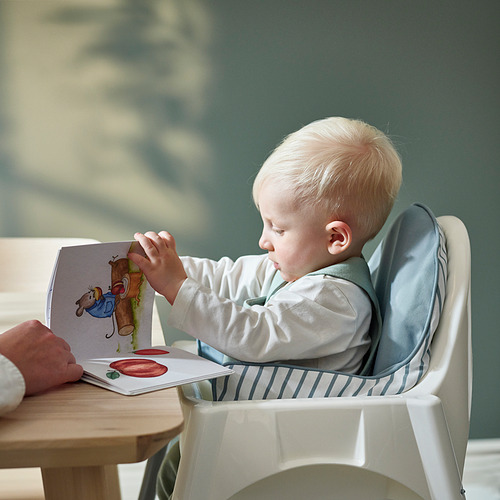 Bebé en silla alta, leyendo con un adulto, sobre un cojín inflable reversible para apoyarse.