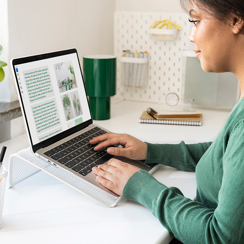 Usando un soporte plateado para computadora portátil DRÖNJÖNS en un escritorio blanco, con lámpara verde, para una mejor postura.