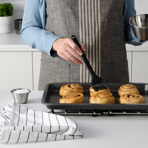 Una persona está cepillando productos horneados con un cepillo de silicona. Una bandeja para hornear y los ingredientes están sobre la mesa.