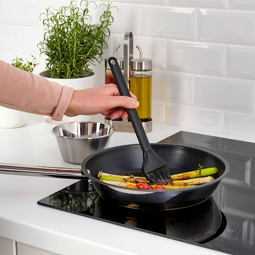 Persona asando verduras con un cepillo GRILLTIDER en estufa negra, con hierbas y aceite en el fondo.