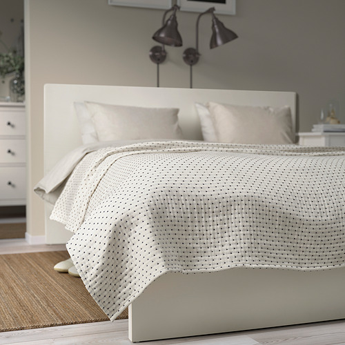 Colcha de color blanco roto con costuras en gris oscuro sobre una cama grande, parte de una configuración de dormitorio minimalista.
