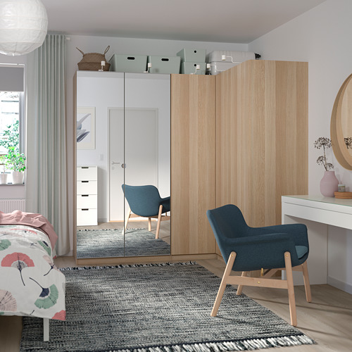 Dormitorio con armario de madera clara, espejo grande, silla azul, escritorio blanco, alfombra y cama con manta de colores.