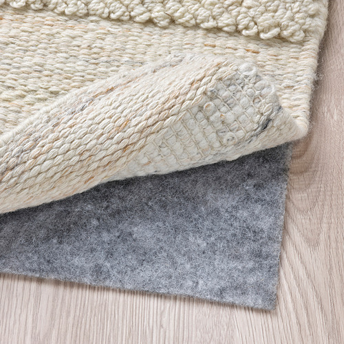 Un primer plano de una alfombra tejida de color beige colocada sobre un suelo de madera clara, con una capa de fieltro gris visible, que muestra su textura y ubicación funcional.