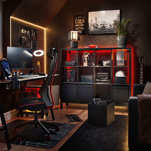 Moderna oficina en casa con estanterías negras KALLAX, luces rojas, PC de sobremesa, silla ergonómica, guitarra y decoración.