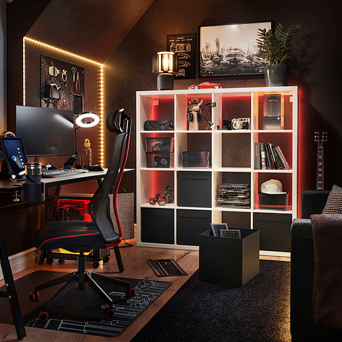 Moderna oficina en casa con silla de escritorio negra y roja, estantería KALLAX blanca con iluminación led, guitarra y equipo tecnológico.