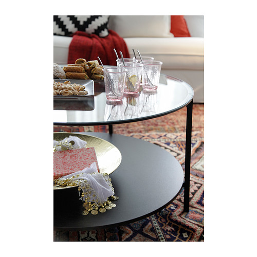 Mesa de centro redonda de cristal con estructura metálica. Sostiene bebidas y bocadillos en la parte superior, con un estante inferior para almacenamiento.