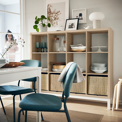 Acogedor comedor con estantes KALLAX que contienen platos, jarrones, libros y plantas. Silla azul, lámpara blanca, ambiente luminoso.