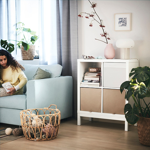 Mujer lee en un sofá azul claro de la estantería KALLAX con cesta de hilo, libros y plantas.