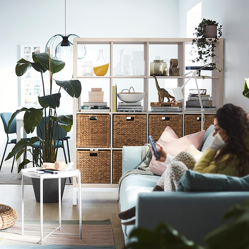 Mujer en sofá azul usando teléfono, estantería KALLAX con plantas, habitación luminosa.