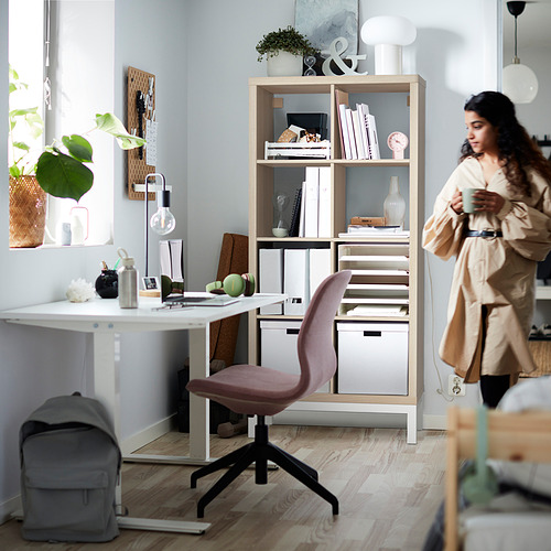 Una mujer está de pie junto a una estantería KALLAX de madera clara con una silla de escritorio rosa, sosteniendo una taza de café en una habitación luminosa y organizada.