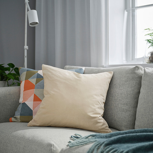 Sala de estar con cojín SANELA beige, almohada multicolor, manta verde, planta, lámpara y ventana con cortina.