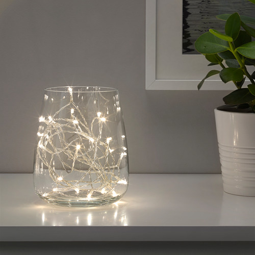 Jarrón de vidrio transparente con luces led blancas cálidas que proyectan un brillo festivo en un estante blanco.