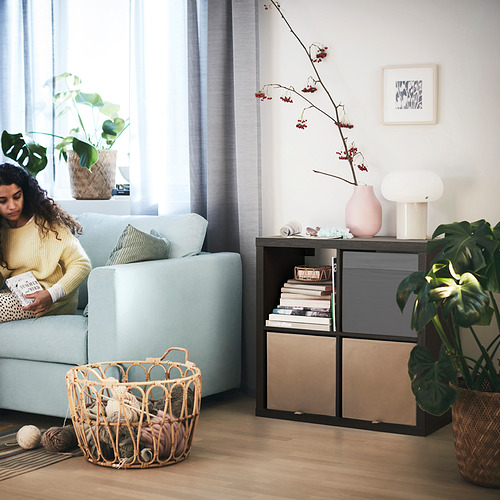 Sala de estar con estantería KALLAX y sofá azul claro. Mujer leyendo, cesta de hilo y plantas.