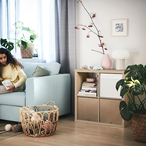 Mujer leyendo en sofá azul, estante KALLAX con libros y flores, plantas, cesta de hilo, cortinas azules, pintura mural.