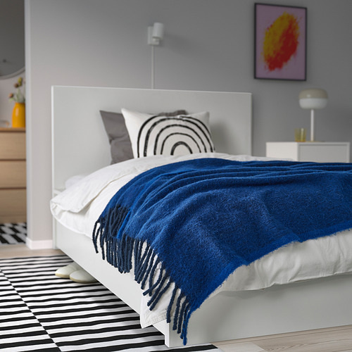 Dormitorio moderno con cama blanca de líneas limpias, manta con flecos azules, alfombra estampada, arte colorido y mesita de noche minimalista. Acogedor, acogedor y elegante.