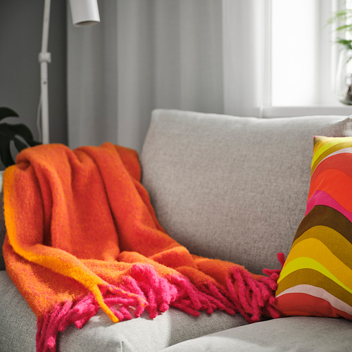 Manta KLIPPOXEL rosa brillante/naranja en el sofá, suave y esponjosa, con extremos de flecos, cerca de una ventana grande, cubre parte del cojín gris.