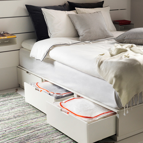 Dormitorio con bolsa de lino PÄRKLA debajo de la cama, cremallera roja, almohadas a rayas, libro, lámpara.