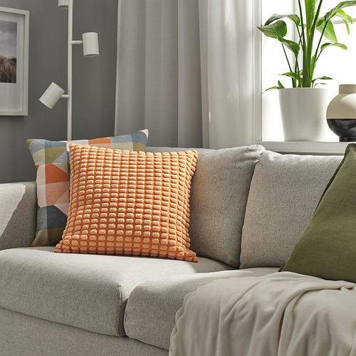 Sofá gris moderno con gran cojín naranja, manta verde, planta, cuadro, iluminación acogedora.