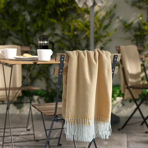 Manta beige-amarilla HOLMVI con flecos doblados sobre la silla, más grande que tazas de café y platos sobre la mesa.