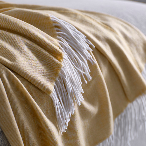Manta doblada de color beige-amarillo con bordes de flecos sobre una superficie blanca. Textura suave, poliéster reciclado.